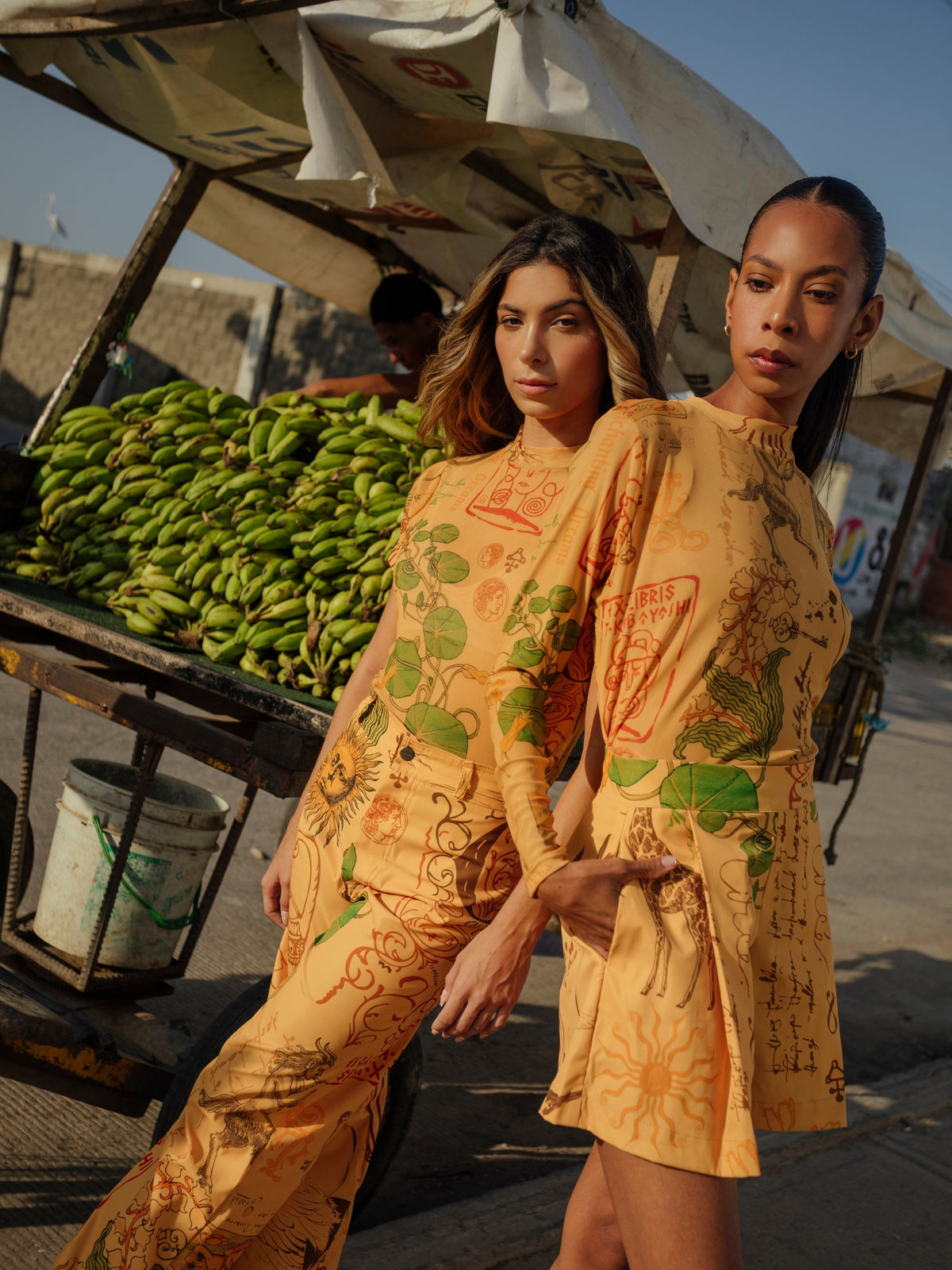 Two models in the Circe Trofo ochre printed set beside a banana stall
