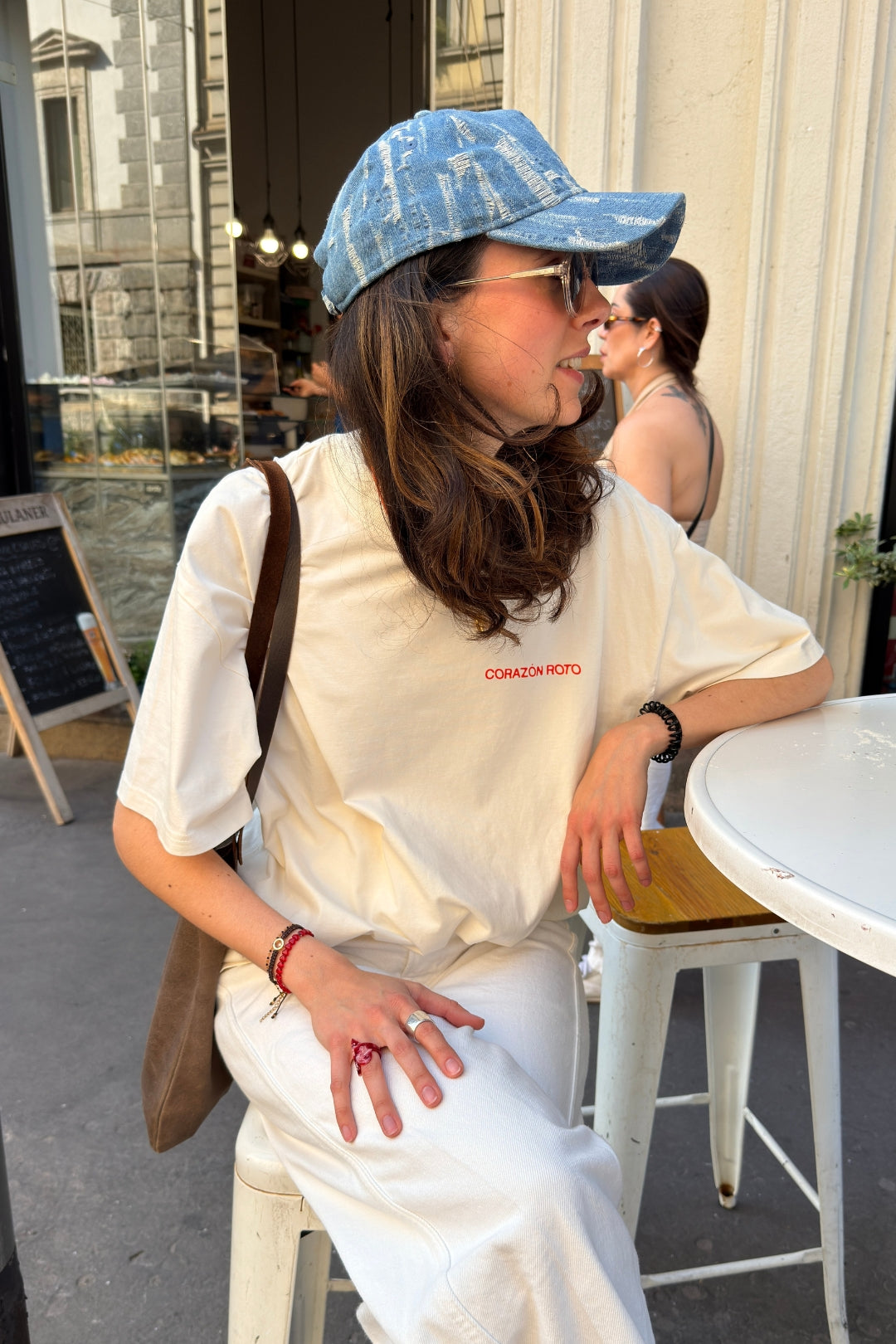 Woman in Limited Edition CR T-shirt cream oversized graphic tee with blue cap cafe setting - Corazon Roto