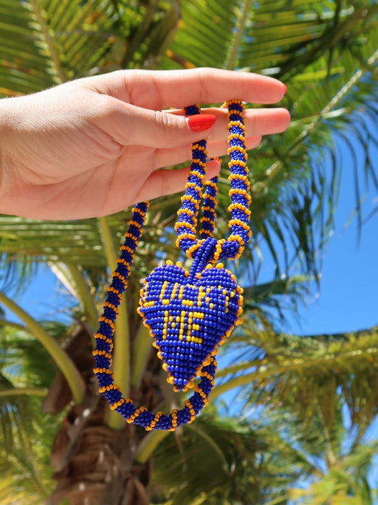 Long Lucky Me beaded heart necklace by Pajara Pinta held in a hand under palm trees