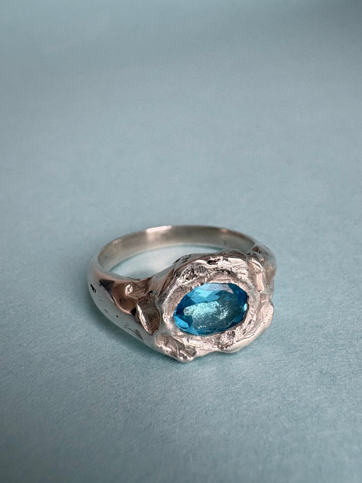 STUDIO MUREA Azul Profundo Ring, a blue topaz in a molten-look silver setting, on a pale blue background